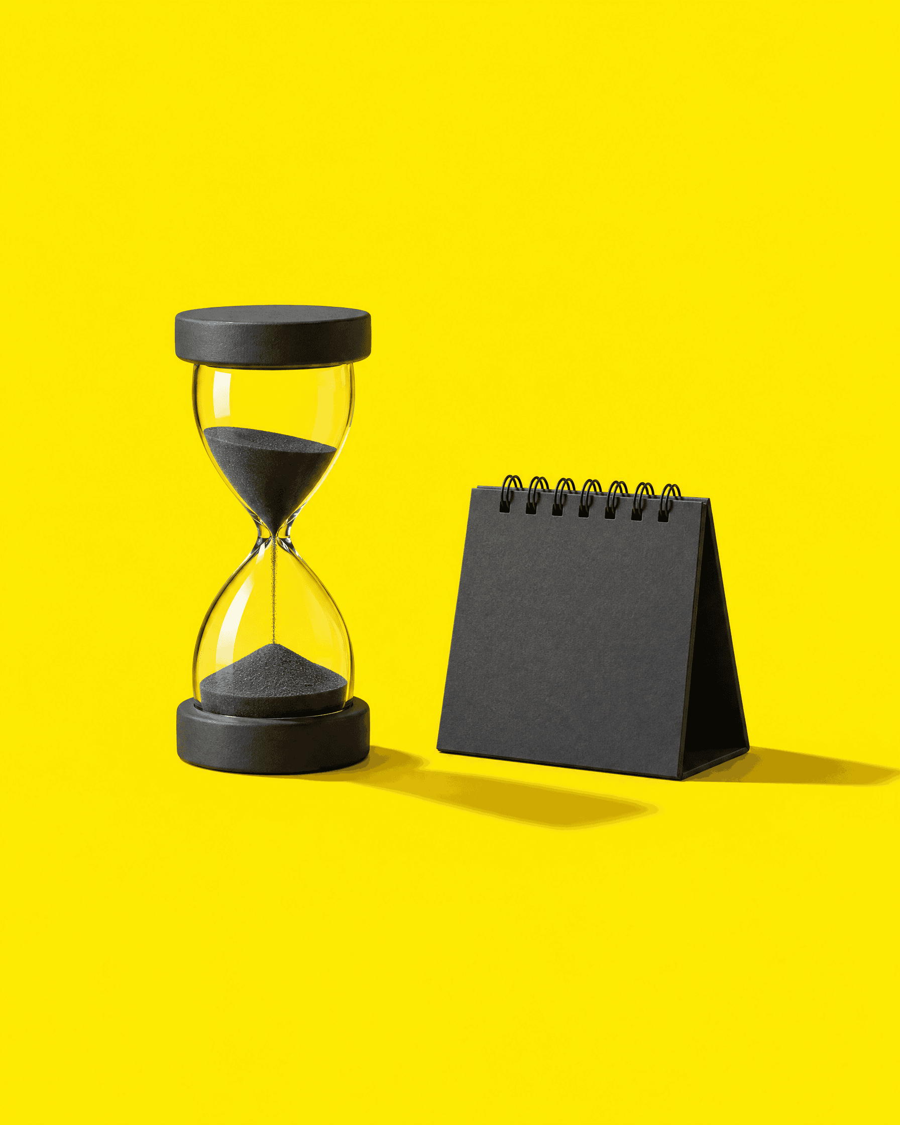 Eine anthrazitfarbene Sanduhr neben einem kleinen Tischkalender auf gelbem Grund